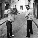 Two men stand on a narrow cobblestone street in a city. One has a bag slung over his shoulder and is looking back, while the other, holding items in his arms, smiles at the camera. The image is in black and white.